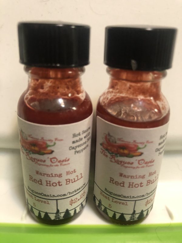 Two bottles of Rogues Oasis Red Hot Bull hot sauce with spicy red liquid, labeled with heat level 5.