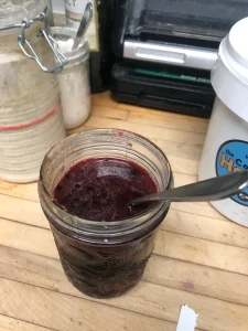 Vibrant blackberry shrub mix in a glass jar with a spoon, surrounded by rustic kitchen elements.